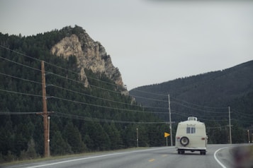 A vintage white camper van travels on a winding highway flanked by dense evergreen forests and a rocky mountain. Overhead, power lines run parallel to the road. The sky is overcast, casting a subdued light over the scene.