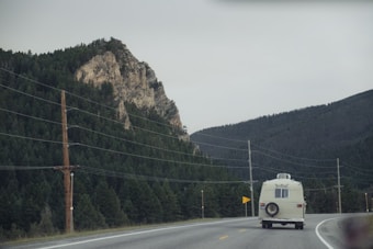 A vintage white camper van travels on a winding highway flanked by dense evergreen forests and a rocky mountain. Overhead, power lines run parallel to the road. The sky is overcast, casting a subdued light over the scene.