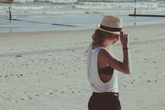 A model wearing a breezy linen shirt and wide-brimmed hat walking along the shore.