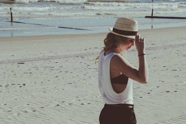 A model wearing a breezy linen shirt and wide-brimmed hat walking along the shore.