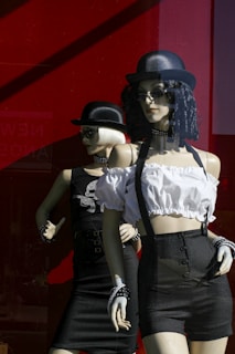 Two mannequins dressed in stylish clothing are positioned in front of a bright red backdrop. The one in the foreground wears a white cropped blouse with suspenders and black high-waisted shorts, accessorized with a black choker and hat. The other mannequin is dressed in a black outfit with patterned embellishments, also wearing a black hat and sunglasses. The setting appears to be a store display window.