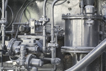 A complex assembly of industrial machinery components with numerous metal pipes, valves, and connectors. The surface has a metallic texture with a greyish color, and the equipment appears to be well-maintained.