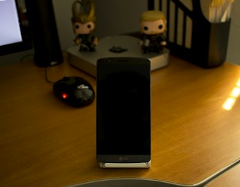 A wooden desk holds a smartphone with the LG logo on its front, positioned in the center. To the left, a black computer mouse with a glowing red light is visible. In the background, two figurines resembling toys are placed on top of a device that looks like a Mac Mini. A pencil holder containing a few writing instruments is seen on the right side of the desk. Lighting creates a subtle highlight on the desk's surface.