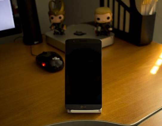 A wooden desk holds a smartphone with the LG logo on its front, positioned in the center. To the left, a black computer mouse with a glowing red light is visible. In the background, two figurines resembling toys are placed on top of a device that looks like a Mac Mini. A pencil holder containing a few writing instruments is seen on the right side of the desk. Lighting creates a subtle highlight on the desk's surface.