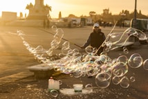 A person creates large, iridescent soap bubbles using two long sticks with strings attached. The setting is an outdoor urban area during sunset, with warm golden sunlight spreading across the scene. Various people and structures are visible in the background, slightly out of focus.