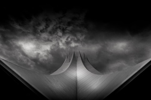 A dramatic, dark and moody image featuring a futuristic architectural structure with sharp, angular, upward-pointing peaks. The background showcases ominous, swirling clouds that enhance the sense of tension and mystery.