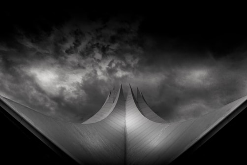 A dramatic, dark and moody image featuring a futuristic architectural structure with sharp, angular, upward-pointing peaks. The background showcases ominous, swirling clouds that enhance the sense of tension and mystery.