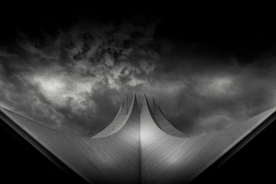 A dramatic, dark and moody image featuring a futuristic architectural structure with sharp, angular, upward-pointing peaks. The background showcases ominous, swirling clouds that enhance the sense of tension and mystery.