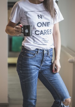 A person is wearing a white T-shirt with the text 'No One Cares' printed on it, along with blue jeans. The person is holding a black mug with a logo. The setting appears to be indoors with a neutral-colored background.