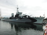A large naval warship is docked at a harbor, displaying a camouflage pattern with the designation 'H34' prominently visible on its side. The ship has several turrets and antenna structures on its deck, and appears to be stationary. The water around the ship is calm, reflecting parts of the vessel. The sky is overcast, giving the scene a muted and subdued atmosphere.