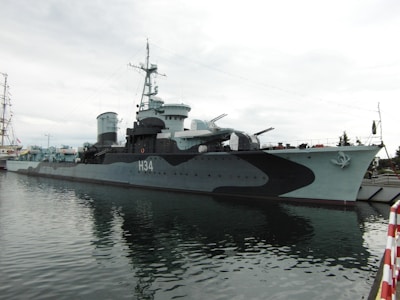 A large naval warship is docked at a harbor, displaying a camouflage pattern with the designation 'H34' prominently visible on its side. The ship has several turrets and antenna structures on its deck, and appears to be stationary. The water around the ship is calm, reflecting parts of the vessel. The sky is overcast, giving the scene a muted and subdued atmosphere.