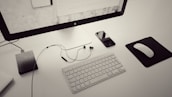 A minimalist workspace featuring a large monitor on a stand, a wireless keyboard, and a mouse on a black mousepad. To the left, there is a textured external hard drive connected via a cable, while a smartphone with a reflective screen and earbuds rests beside the keyboard. The color scheme is primarily monochrome with a sleek, modern aesthetic.