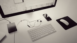 A minimalist workspace featuring a large monitor on a stand, a wireless keyboard, and a mouse on a black mousepad. To the left, there is a textured external hard drive connected via a cable, while a smartphone with a reflective screen and earbuds rests beside the keyboard. The color scheme is primarily monochrome with a sleek, modern aesthetic.