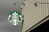 A large circular green and white logo featuring a mermaid is displayed on the side of a building. The building has vertical ridges and extends upwards with a modern industrial design. Overhead lights are attached to the structure, enhancing visibility of the logo.