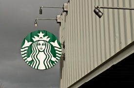 A large circular green and white logo featuring a mermaid is displayed on the side of a building. The building has vertical ridges and extends upwards with a modern industrial design. Overhead lights are attached to the structure, enhancing visibility of the logo.