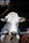 A close-up view of a white and black cow with a numbered tag on its ear, positioned behind a metal fence. The background is dimly lit, suggesting an indoor area with wooden beams above.