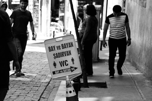 A black and white photograph of a street scene with people walking on a sidewalk. The focus is on a sign indicating public restrooms for men and women, with the words 'BAY ve BAYAN ŞADIRVAN WC' and directional arrows. The cobblestone path and worn walls add texture to the urban setting.