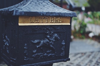 An ornate black mailbox with a gold plaque displaying the word 'LETTERS'. The top features a decorative design and there is an intricate pattern with a horseman on the body of the box. Raindrops are visible on the surface, suggesting a rainy day. The background is blurred, focusing attention on the mailbox.
