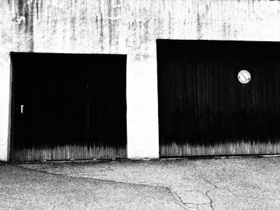 Two garage doors are set in a textured wall, one with a handle and the other featuring a circular no-parking sign. The ground in front is cracked and rough.