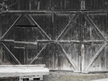A large wooden barn door with a rustic appearance and visible signs of weathering. The doors are made of dark, old wood with diagonal planks forming geometric patterns. Part of the door is slightly ajar. In front of the door is a simple wooden bench on a small pallet.