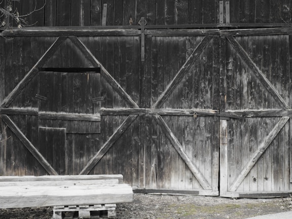 A large wooden barn door with a rustic appearance and visible signs of weathering. The doors are made of dark, old wood with diagonal planks forming geometric patterns. Part of the door is slightly ajar. In front of the door is a simple wooden bench on a small pallet.