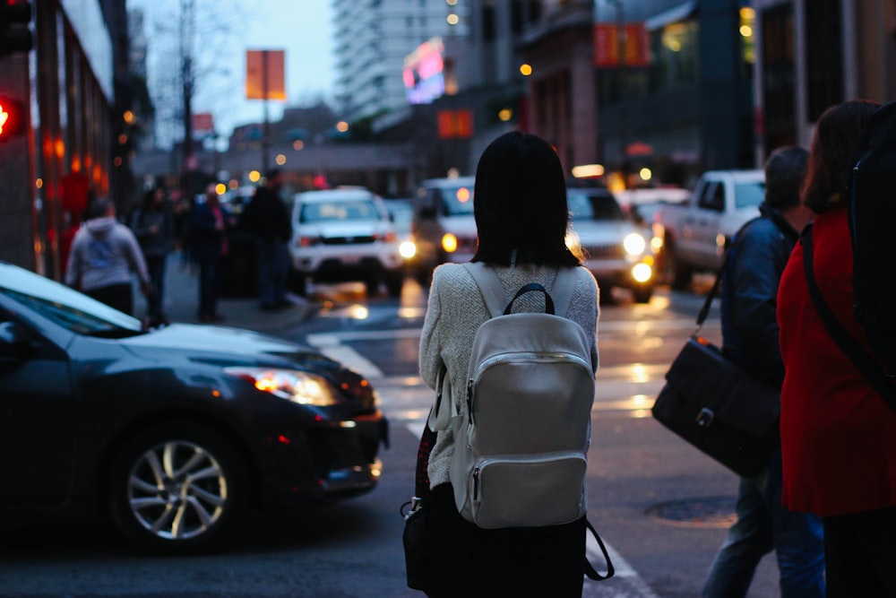 woman wearing grey shirt and white backpack facing car on street