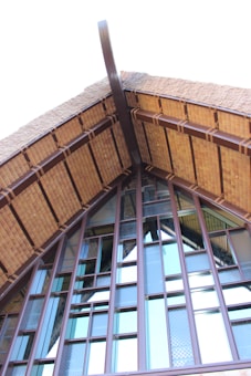 A close-up of a building's roof structure, featuring a steep gable design with wooden beams and textured glass panels. The architecture highlights a mixture of traditional and modern elements, with prominent use of natural materials.