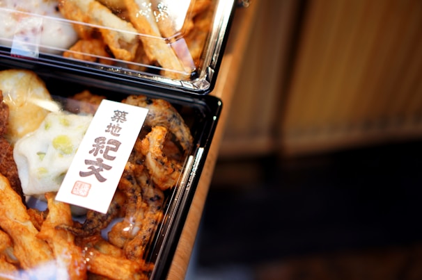 A close-up view of packaged food items in black plastic containers, likely containing various types of deep-fried snacks. There is a label with Japanese characters, possibly indicating the brand or type of snacks.