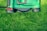A lawn mower is positioned on a patch of lush, green grass, showcasing the machine's green and black exterior with a red lever on the side.