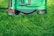 A lawn mower is positioned on a patch of lush, green grass, showcasing the machine's green and black exterior with a red lever on the side.