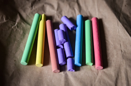 Several sticks of colored chalk are arranged in a row on a textured, brown surface. The chalk pieces are vibrant and include colors such as green, yellow, pink, purple, and blue.