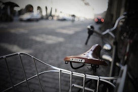 A close-up view of a vintage-style brown leather Brooks bicycle saddle and part of a metal wire basket on a bike. The background features a blurred urban street scene, with cars and a structure with arches, adding depth to the composition.