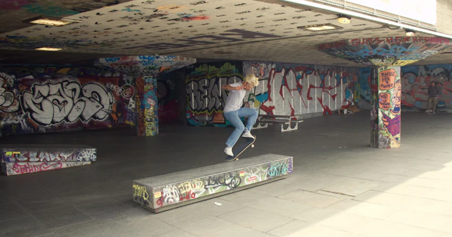 A skateboarder performing a kickflip on a sunny urban skatepark with graffiti walls.