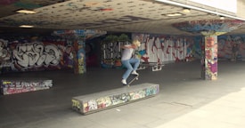 A skateboarder performing a trick on a graffiti-covered concrete platform in an urban skate park. The area is decorated with vibrant street art on the walls and pillars, creating a colorful and energetic backdrop. The skateboarder is mid-action, showcasing skill and balance.