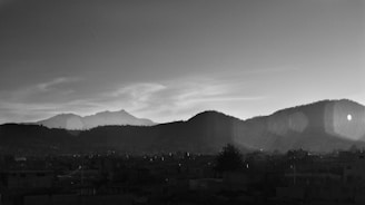 A black and white landscape featuring silhouetted mountains against a sky with scattered clouds. The foreground shows a cityscape with buildings in shadow, and some lens flare is visible in the top right corner.
