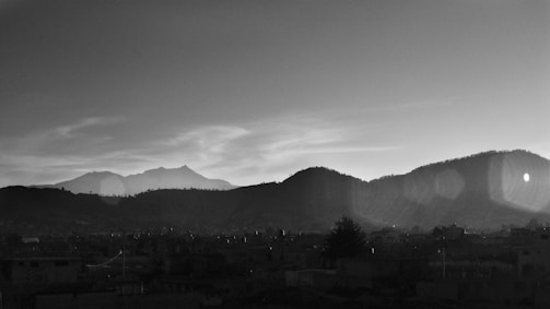A black and white landscape featuring silhouetted mountains against a sky with scattered clouds. The foreground shows a cityscape with buildings in shadow, and some lens flare is visible in the top right corner.