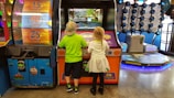 Two children stand in front of an arcade game, each actively engaged with the controls. The environment is colorful and lively, with various game machines and ticket displays in the background. The floor is shiny, reflecting the overhead lights.