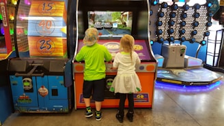 Two children stand in front of an arcade game, each actively engaged with the controls. The environment is colorful and lively, with various game machines and ticket displays in the background. The floor is shiny, reflecting the overhead lights.
