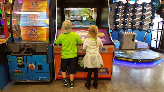 Two children stand in front of an arcade game, each actively engaged with the controls. The environment is colorful and lively, with various game machines and ticket displays in the background. The floor is shiny, reflecting the overhead lights.