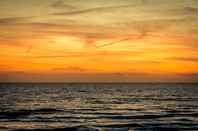 A serene ocean view at sunset with orange and yellow hues filling the sky. Gentle waves ripple across the surface of the water, creating a calm and peaceful atmosphere.