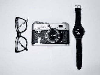black and silver camera; eyeglasses with black frames; black analog watch