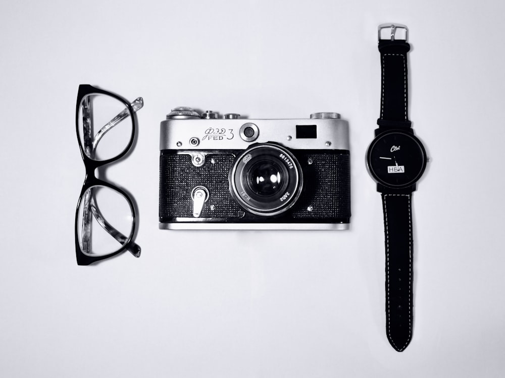 black and silver camera; eyeglasses with black frames; black analog watch