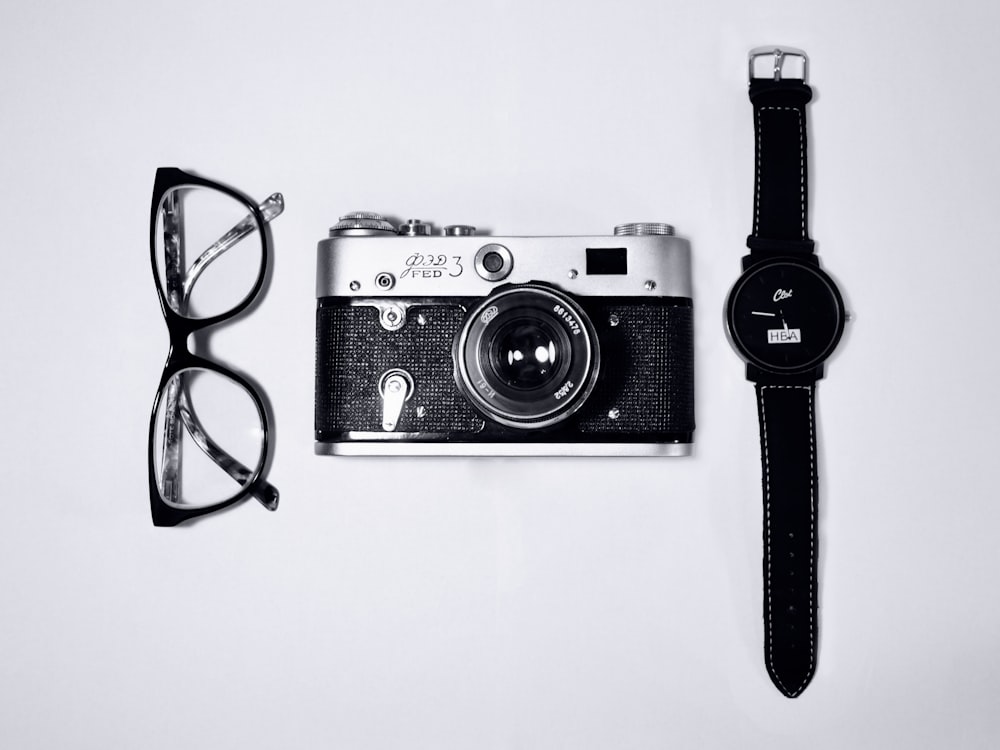câmera preta e prata; óculos com armações pretas; relógio analógico preto