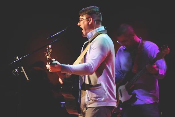 A musician is passionately singing into a microphone while playing an acoustic guitar. Another person is slightly behind him, playing an electric bass. The setting appears to be a dimly lit stage with warm, atmospheric lighting.