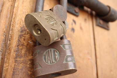 Two vintage Yale padlocks are attached to a wooden door. The locks are made of metal with a bronze hue, and the Yale logo is prominently displayed on each lock. The background consists of a textured wooden surface with warm tones.