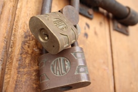 Two vintage Yale padlocks are attached to a wooden door. The locks are made of metal with a bronze hue, and the Yale logo is prominently displayed on each lock. The background consists of a textured wooden surface with warm tones.