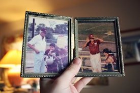 A hand holds a double-picture frame containing two vintage photographs. The left photo depicts an older man wearing a hat and light-colored clothing, standing with two young boys in sports uniforms. The right photo shows a man in a maroon hat and shirt, saluting or shielding his eyes from the sun, standing on a sports field.