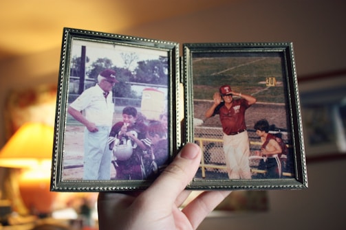 A hand holds a double-picture frame containing two vintage photographs. The left photo depicts an older man wearing a hat and light-colored clothing, standing with two young boys in sports uniforms. The right photo shows a man in a maroon hat and shirt, saluting or shielding his eyes from the sun, standing on a sports field.