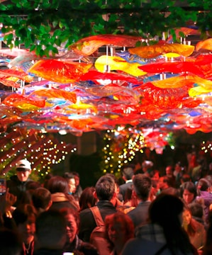 A crowded indoor setting with vibrant, colorful decorations hanging from the ceiling, resembling abstract or artistic glass shapes in reds, yellows, and blues. People gather beneath these decorations, with warm, ambient lighting contributing to a lively atmosphere. Green leaves are visible at the top, possibly as part of the decoration. Strings of small lights add a festive touch.