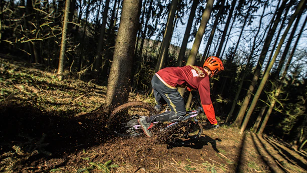 A person wearing a red jacket and orange helmet is mountain biking through a forest. The bike kicks up dirt as the rider navigates a sharp turn on a narrow trail. Tall trees surround the path, and sunlight filters through the branches, creating a dynamic and adventurous scene.
